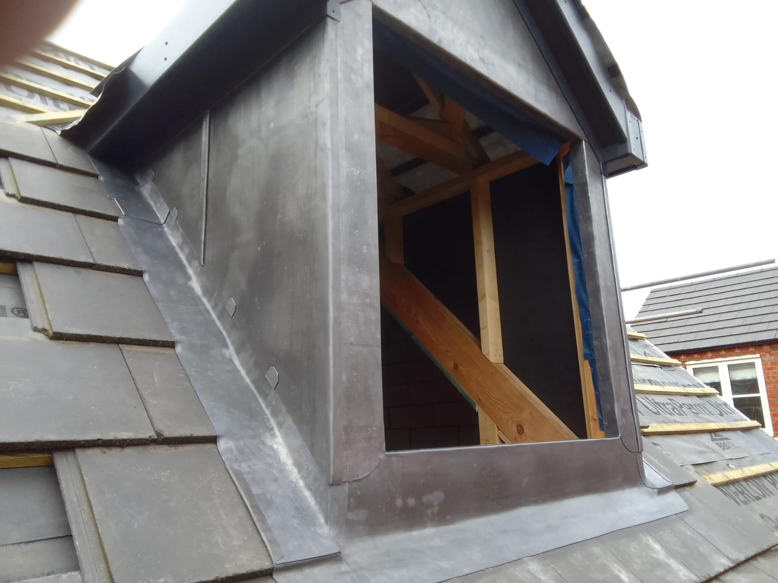 a gray slate roof with a dormer under construction. the structure has exposed wooden beams and metal flashing, conveying a work in progress feel