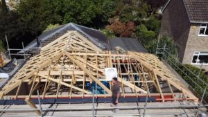 a man walks on scaffolding in front of a partially constructed wooden roof framework of a house extension, surrounded by greenery and nearby homes