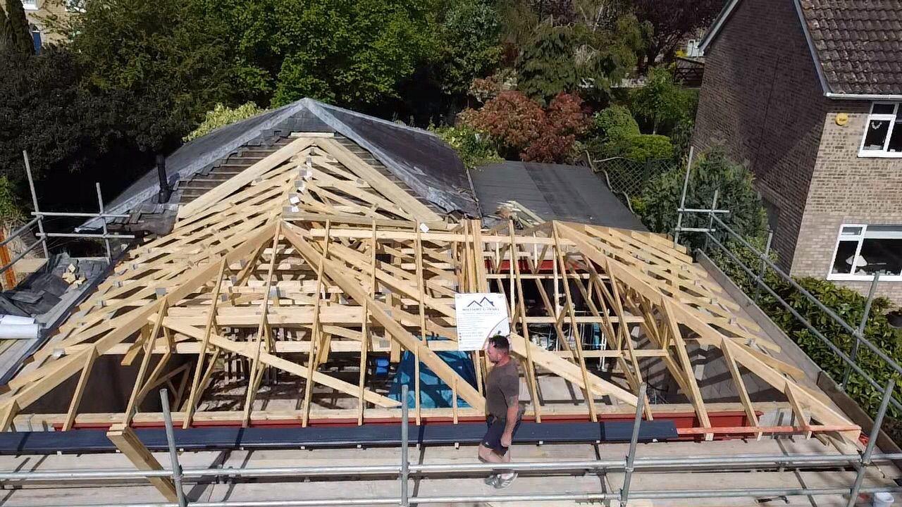 a man walks on scaffolding in front of a partially constructed wooden roof framework of a house extension, surrounded by greenery and nearby homes
