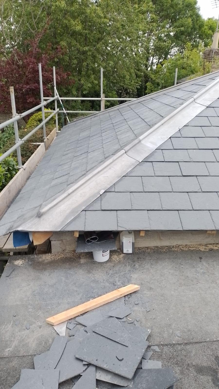 a newly tiled roof with gray slate shingles, scaffolding on the left, trees in the background, and a few scattered tiles on the foreground rooftop
