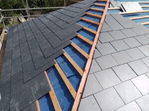 a roof under construction with gray slate tiles partially installed. wooden beams and blue underlayment are visible. the setting appears calm and orderly
