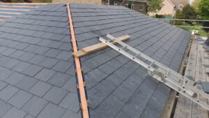 a slate roof under construction, showing a neat pattern of gray tiles. a ladder rests on a wooden beam across the roof, with houses visible in the background