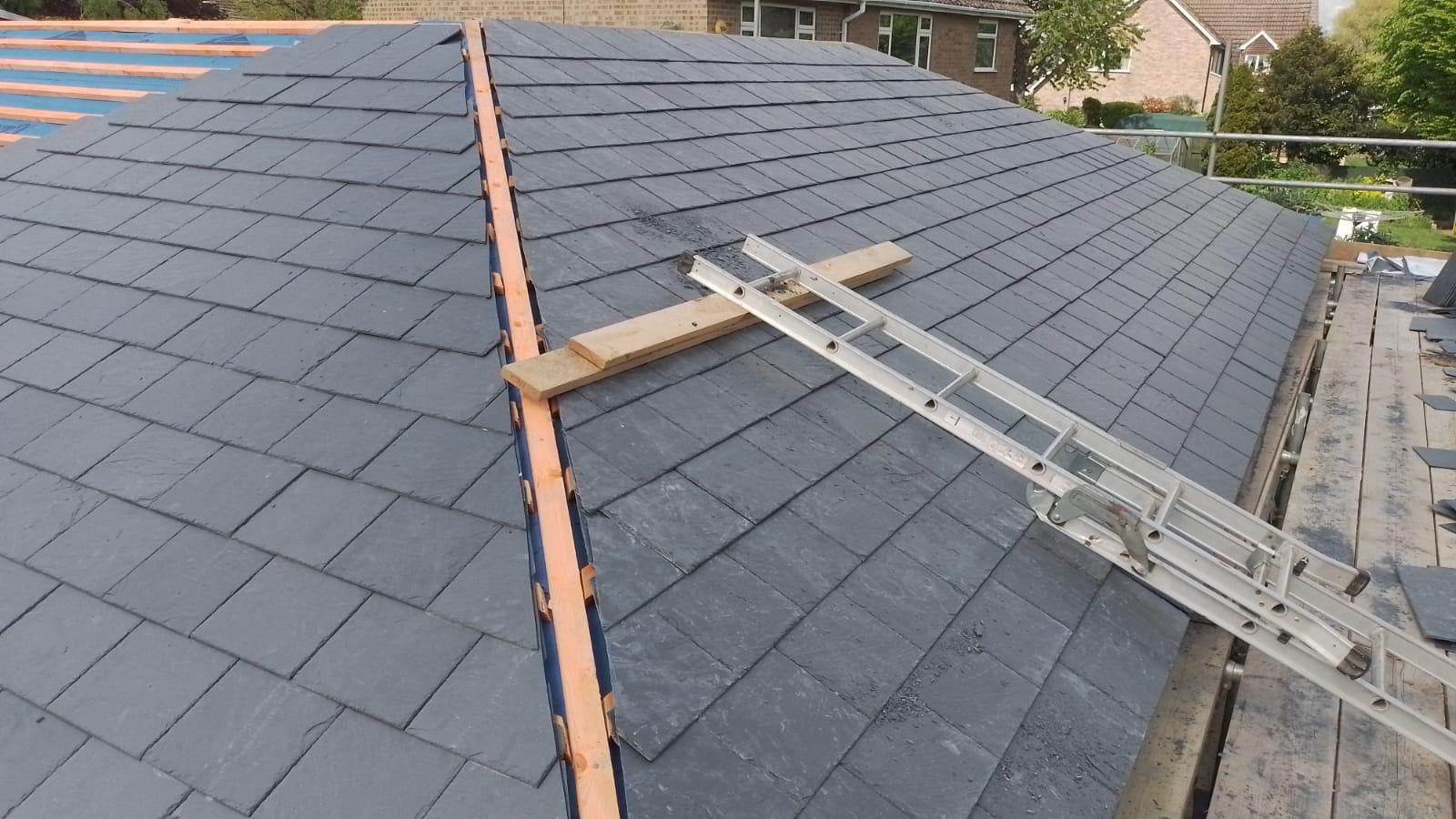 a slate roof under construction, showing a neat pattern of gray tiles. a ladder rests on a wooden beam across the roof, with houses visible in the background