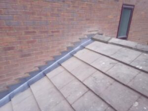 brick wall meeting a tiled roof with step flashing in between for waterproofing. the roof tiles are slightly weathered, and a small window is visible above