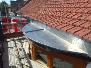 red tiled roof with new lead flashing, viewed from scaffolding. overcast sky and adjacent buildings visible. construction work in progress