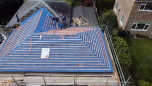 two workers install a blue roof with wooden beams on a building, surrounded by greenery. scaffolding is visible, indicating ongoing construction