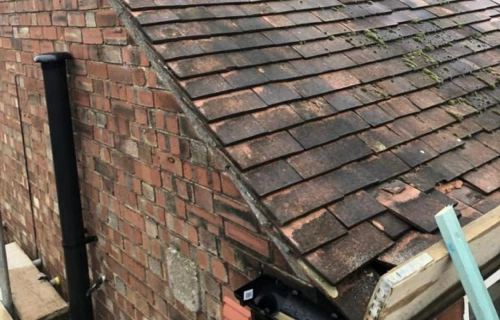 Home worker sitting on a steep brick tiled roof, wearing a safety vest. the brick chimney and surrounding buildings suggest a residential area. overcast sky